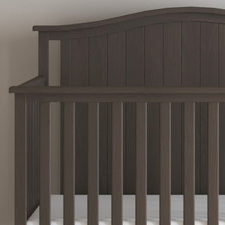 The Child Craft Hampton Arch Top 4-in-1 Convertible Crib in Dapper Grey features vertical slats, a curved headboard, and an adjustable mattress support with a white mattress, set against a light-colored wall.