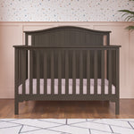 The Child Craft Hampton Arch Top 4-in-1 Convertible Crib in Dapper Grey, with vertical slats and a pale pink mattress, stands on a wooden floor. Behind it, a wall is painted light pink with a white top portion featuring colorful dots.