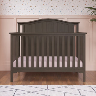 The Child Craft Hampton Arch Top 4-in-1 Convertible Crib in Dapper Grey, with vertical slats and a pale pink mattress, stands on a wooden floor. Behind it, a wall is painted light pink with a white top portion featuring colorful dots.