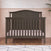 The Child Craft Hampton Arch Top 4-in-1 Convertible Crib in Dapper Grey, with vertical slats and a pale pink mattress, stands on a wooden floor. Behind it, a wall is painted light pink with a white top portion featuring colorful dots.