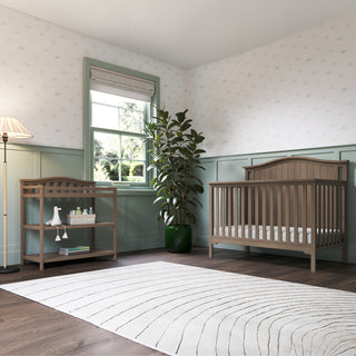 A cozy nursery features a wooden crib, Child Craft Arch Top Baby Changing Table in Dusty Heather with storage, large potted plant, window view, lamp, and patterned white rug on dark wood floors. Walls have light green wainscoting and wallpaper.