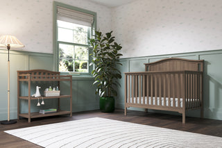 A cozy nursery with a wooden crib, Child Craft's Arch Top Baby Changing Table in Dusty Heather for storage, a tall plant in a green pot, a floor lamp, natural window light, and playful patterned wallpaper on green and white walls.