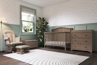 A cozy nursery with a wooden crib, dresser, plant, and Child Craft Cozy Glider and Ottoman in Dusty Heather. Pale green wainscoting, a sunlit window, and a white rug with curved lines complete the space.