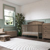A softly lit nursery with green wainscoting features the Child Craft Hampton Arch Top 4-in-1 Convertible Crib in Dusty Heather, matching dresser, rocking chair with ottoman, toy chest, potted plant, blinds, and a curved-line white rug on dark wood floors.