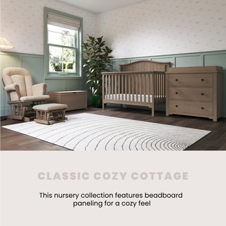 A cozy nursery with green beadboard paneling, a Child Craft Hampton Arch Top 4-in-1 Convertible Crib in Dusty Heather, dresser, rocking chair with ottoman, and large rug on dark wood floors; sunlight streams through two windows.