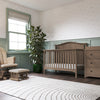 A cozy nursery with the Child Craft Hampton Arch Top 4-in-1 Convertible Crib in Dusty Heather, matching dresser, rocking chair and ottoman, tall plant, floor lamp, pale green and white patterned walls, a white rug, and dark wood floors.