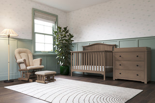 A cozy nursery with the Child Craft Hampton Arch Top 4-in-1 Convertible Crib in Dusty Heather, matching dresser, rocking chair and ottoman, tall plant, floor lamp, pale green and white patterned walls, a white rug, and dark wood floors.