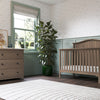 A cozy nursery features the Child Craft Hampton Arch Top 4-in-1 Convertible Crib in Dusty Heather, a matching dresser with framed art and a floor lamp, a tall plant, Roman shade window, patterned wallpaper, and a round white rug on dark wood floors.