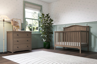 A cozy nursery features the Child Craft Hampton Arch Top 4-in-1 Convertible Crib in Dusty Heather, a matching dresser with framed art and a floor lamp, a tall plant, Roman shade window, patterned wallpaper, and a round white rug on dark wood floors.