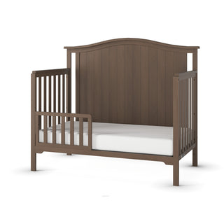 The Child Craft F09501 Toddler Guard Rail in Dusty Heather features a high arched back, vertical slats, a low guard rail for safety, and is shown with a white mattress—ideal for easy crib conversion.