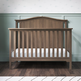 The Child Craft Hampton Arch Top 4-in-1 Convertible Crib in Dusty Heather stands on a dark wood floor against green paneled walls with cloud-print wallpaper. The crib features vertical slats, a curved headboard, and comes with a white mattress.