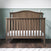The Child Craft Hampton Arch Top 4-in-1 Convertible Crib in Dusty Heather stands on a dark wood floor against green paneled walls with cloud-print wallpaper. The crib features vertical slats, a curved headboard, and comes with a white mattress.