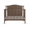 The Child Craft Hampton Arch Top 4-in-1 Convertible Crib in Dusty Heather features vertical slats, a curved headboard, a white mattress, and a simple classic design, shown from the front.