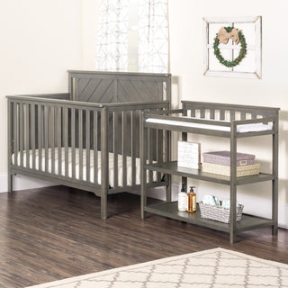 Child Craft Flat Top Baby Changing Table in Dapper Gray in a nursery with wooden floors, beige rug, wall mirror, and framed wreath. Includes changing pad, baskets, towels, and storage for baby products.
