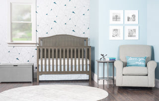 A modern nursery featuring the Child Craft Harmony 33" Kids Toy Box in cool gray, patterned wallpaper, a round rug, a gray rocking chair with a "love you more" pillow, side table, wall art, and natural light from a window.