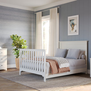 A cozy, modern bedroom featuring a Child Craft F06401 full-size bed with brushed cotton bed rails, gray bedding, nightstand with lamp, mirrored dresser with teddy bear, potted plant, and blue-gray walls lit by window light.