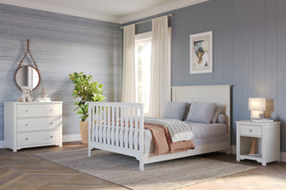 A cozy, modern bedroom featuring a Child Craft F06401 full-size bed with brushed cotton bed rails, gray bedding, nightstand with lamp, mirrored dresser with teddy bear, potted plant, and blue-gray walls lit by window light.