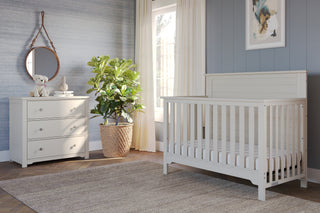 A bright, modern nursery with a white crib, Child Craft Harmony 3-Drawer Dresser with Changing Table Topper in Brushed Cotton, a mirror above, stuffed dog, basket plant, neutral curtains, soft rug, and framed abstract art on a blue-gray wall.