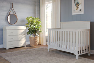 A bright, modern nursery with a white crib, Child Craft Harmony 3-Drawer Dresser with Changing Table Topper in Brushed Cotton, round mirror above, large potted plant, gray walls, sheer-curtained window, beige rug, and framed abstract art.