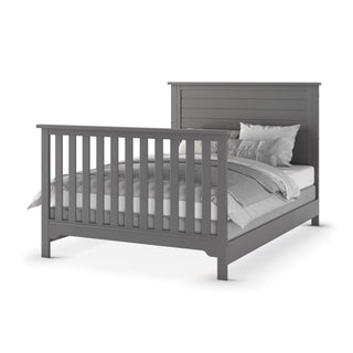 The Child Craft F06401 Full-Size Bed Rails in Brushed Pebble are shown on a gray wooden bed frame with vertical slats, dressed in white and gray bedding with pillows and a quilt, all set against a white background.