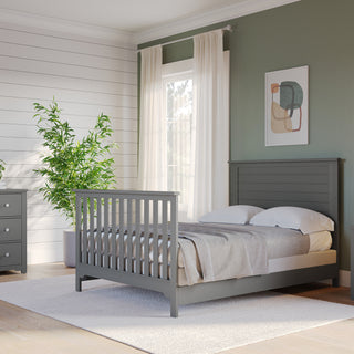 A modern bedroom featuring the Child Craft F06401 Full-Size Bed Rails in Brushed Pebble on a gray bed, with a matching nightstand and dresser, round mirror, potted plant, white-curtained window, and art on the green accent wall.