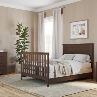 A cozy bedroom features a pine wood bed, nightstand, dresser, light walls, large plant, round mirror, framed art, and soft curtains over two windows. Neutral bedding and rug add warmth. Child Craft F06401 Full-Size Bed Rails in Brushed Truffle offer style and support.