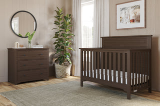 A cozy nursery with a dark wood crib, the Child Craft Harmony 3-Drawer Dresser with Changing Table Topper in Brushed Truffle, a circular mirror, potted plant, framed wall art, woven rug on wooden floors, and sunlight through white-curtained windows.