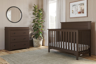 A cozy nursery features a dark wooden crib, the Child Craft Harmony 3-Drawer Dresser with Changing Table Topper in Brushed Truffle, a round wall mirror, potted plant, light curtains, framed art on beige paneled walls, and a woven rug on the wooden floor.