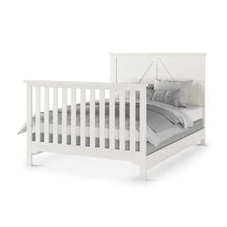 The Child Craft Woodland 4-in-1 Convertible Crib in Brushed Cotton features a white wooden frame with slatted headboard and footboard, and is shown neatly made with gray and white bedding and pillows.