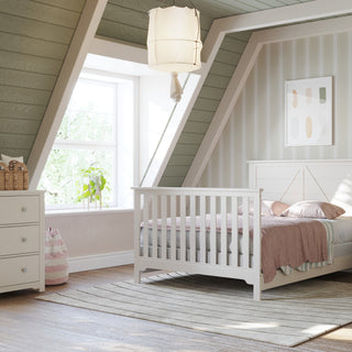 A cozy attic children's bedroom with slanted green walls, white furniture including a bed with Child Craft F06401 Full-Size Bed Rails in Brushed Cotton, dresser, nightstand, stuffed animals, a sunlit window, and soft neutral decor.