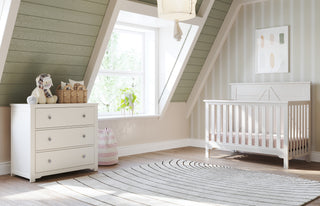 A cozy nursery features a white crib, the Child Craft Harmony 3-Drawer Dresser with Changing Table Topper in Brushed Cotton, stuffed toys, a striped laundry basket, and a patterned round rug on wooden floors, with soft green and beige walls for warmth.