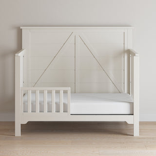 The Child Craft Woodland 4-in-1 Convertible Crib in Brushed Cotton features a paneled headboard and partial side rails, with a simple modern farmhouse design, shown against a light wall and floor.