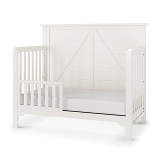 The Child Craft Woodland 4-in-1 Convertible Crib in Brushed Cotton features a high paneled back, open slatted sides, partial side rail, and a fitted, low-to-ground mattress in a simple modern farmhouse style.