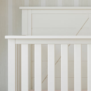A close-up of the Child Craft Woodland 4-in-1 Convertible Crib in Brushed Cotton, featuring white wooden vertical slats, is set against a wall with light-colored striped wallpaper. The crib is empty.