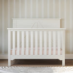 The Child Craft Woodland 4-in-1 Convertible Crib in Brushed Cotton stands against a beige striped wall on a wooden floor, featuring vertical slats, a light pink mattress, and a round black and white patterned rug in front.