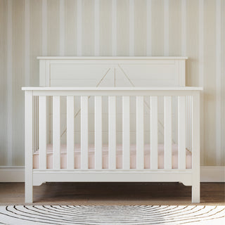 The Child Craft Woodland 4-in-1 Convertible Crib in Brushed Cotton stands against a beige striped wall on a wooden floor, featuring vertical slats, a light pink mattress, and a round black and white patterned rug in front.
