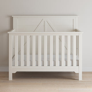 The Child Craft Woodland 4-in-1 Convertible Crib in Brushed Cotton features vertical slats and a decorative headboard in a simple, modern farmhouse style, and is shown on a light wood floor against a plain, light-colored wall.