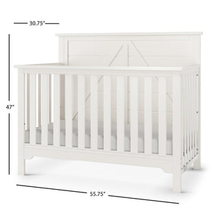 The Child Craft Woodland 4-in-1 Convertible Crib in Brushed Cotton is a white wooden crib with slatted sides, a paneled headboard, and dimensions of 47"H x 30.75"D x 55.75"W.