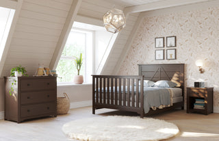 Cozy attic bedroom featuring a dark wood bed, nightstand, and Child Craft Harmony 3-Drawer Dresser with Changing Table Topper in Brushed Truffle, light leaf-patterned walls, round white rug, sunlit window, plants, and wall art above the bed.