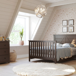 A cozy attic bedroom with sloped ceilings, a large window, and natural light. Features the Child Craft F06401 Full-Size Bed Rails in Brushed Truffle, matching nightstand and dresser, plants, accent wall, and white rug on light wood floors.