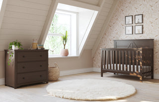 A cozy nursery with a dark wooden crib and the Child Craft Harmony 3-Drawer Dresser with Changing Table Topper in Brushed Truffle, a fluffy white rug, potted plants, and a leaf-patterned accent wall behind the crib. Light streams through a large window.