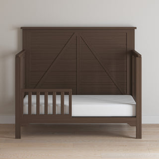The Child Craft F09501 Toddler Guard Rail in Brushed Truffle is a dark brown wooden bed with a high headboard and guard rail, set against a light wall on wood flooring. It features a white mattress and boasts a simple, modern design.