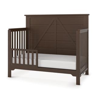 The Child Craft Woodland 4-in-1 Convertible Crib in Brushed Truffle features dark brown pine wood, slatted side rails, a partial safety rail, high paneled headboard, white mattress, and adjustable support for growing toddlers.