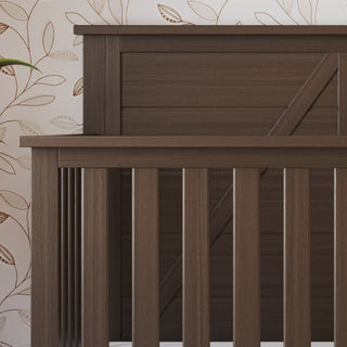 A close-up of the Child Craft Woodland 4-in-1 Convertible Crib in Brushed Truffle shows its dark pine finish and vertical slats, set against a light wallpaper with a brown leaf pattern. An adjustable mattress support is included; a green plant appears on the left.