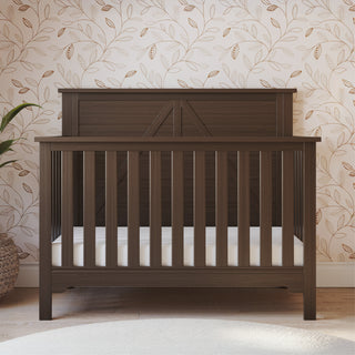 The Child Craft Woodland 4-in-1 Convertible Crib in Brushed Truffle with an adjustable mattress support and white mattress stands on a wooden floor against light floral wallpaper. A green plant is partially visible on the left.