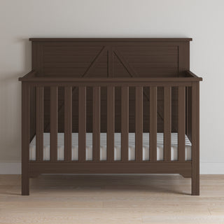 The Child Craft Woodland 4-in-1 Convertible Crib in Brushed Truffle stands on a light wood floor against a white wall, featuring vertical slats, a solid headboard, adjustable mattress support, and a mattress with a white sheet.