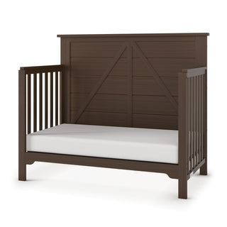 The Child Craft Woodland 4-in-1 Convertible Crib in Brushed Truffle features a high paneled back, open slatted sides, adjustable mattress support, and comes with a white mattress, shown on a plain white background.