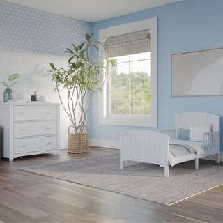 A light blue and white toddler bedroom features the Child Craft Hampton Toddler Bed in matte white with guard rails, a dresser, large potted plant, gray rug, abstract wall art, and a window with a roman shade bringing in natural light.