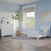 A light blue and white toddler bedroom features the Child Craft Hampton Toddler Bed in matte white with guard rails, a dresser, large potted plant, gray rug, abstract wall art, and a window with a roman shade bringing in natural light.