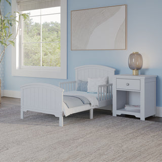 The Child Craft Hampton Toddler Bed in Matte White, featuring white bedding and guard rails, sits low beside a matching nightstand in a light blue room. A large window, abstract art, and a potted plant enhance the cozy, modern vibe.